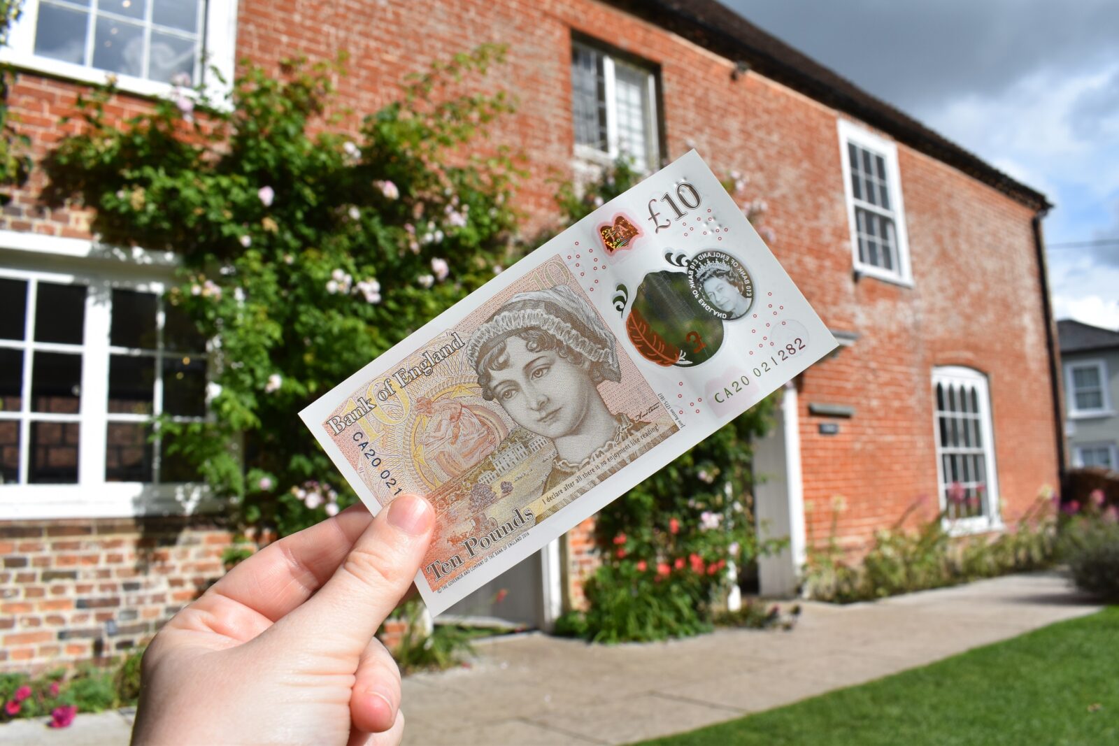 A hand holds up a ten pound note featuring Jane Austen, with Jane Austen's House in the background