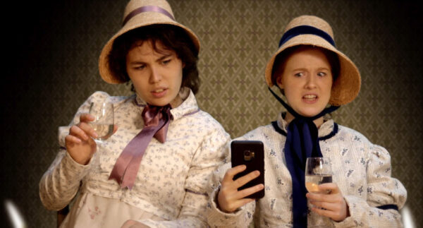 two girls in Regency dress look at a mobile phone in shock